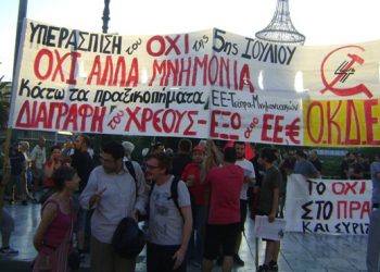 Frente a la bandera de la organización OKDE, en Atenas, en la movilización frente al Parlamento del 22/7, Esther Alcazar de LI de Estado Español y Sedat de IDP de Turquia, junto a un dirigente de OKDE. La bandera dice Defendamos el No del 5 de Julio. No al nuevo Memorando. Contra el crimen de estado de la UE-Tsipras.Cancelacion de la Deuda, Fuera UE y el Euro.