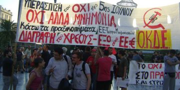 Frente a la bandera de la organización OKDE, en Atenas, en la movilización frente al Parlamento del 22/7, Esther Alcazar de LI de Estado Español y Sedat de IDP de Turquia, junto a un dirigente de OKDE. La bandera dice Defendamos el No del 5 de Julio. No al nuevo Memorando. Contra el crimen de estado de la UE-Tsipras.Cancelacion de la Deuda, Fuera UE y el Euro.