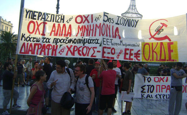 Frente a la bandera de la organización OKDE, en Atenas, en la movilización frente al Parlamento del 22/7, Esther Alcazar de LI de Estado Español y Sedat de IDP de Turquia, junto a un dirigente de OKDE. La bandera dice Defendamos el No del 5 de Julio. No al nuevo Memorando. Contra el crimen de estado de la UE-Tsipras.Cancelacion de la Deuda, Fuera UE y el Euro.